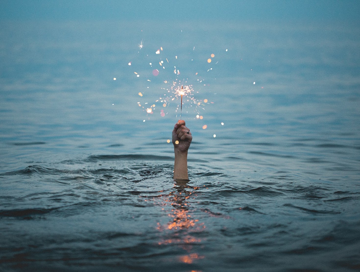 A hand raised above calm water at dusk holding a sparkler